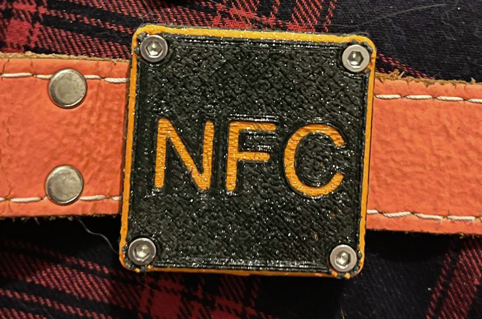 Une photo du support à airtag imprimé en 3D et attaché au collier. Il est noir avec le texte "NFC" orange. Le collier est lui aussi orange.